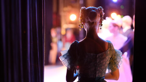 Eine blonde Ballerina in einem türkisen Kleid ist von hinten zu sehen. Sie wartet auf den Moment, in dem sie auf die Bühne tritt. Im Hintergrund sind verschwommen ihre Kolleginnen und Kollegen auf der Bühne zu sehen.