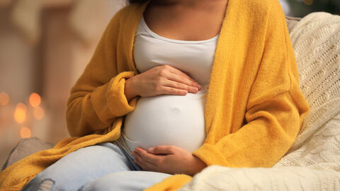 Schwangere Frau sitzt auf einem Stuhl und hält glücklich ihren Bauch fest