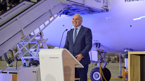 Ein Mann steht an einem Rednerpult. Im Hintergrund ist das Flugzeug.