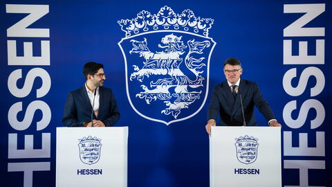 Der stellvertretende Ministerpräsident, Wirtschaftsminister Kaweh Mansoori steht hinter einem Rednerpult links und rechts steht Ministerpräsident Boris Rhein ebenfalls hinter einem Rednerpult. Hinter ihnen ist eine dunkelblaue Wand aufgebaut mit dem weißen Hessen Wappen. An den Seiten steht in weißer Schrift "Hessen"