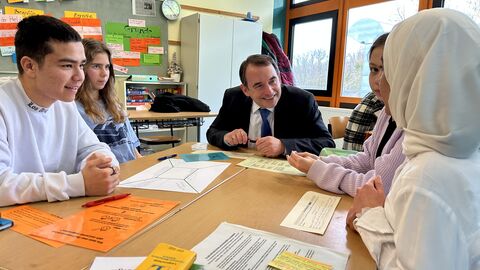 Kultusminister Lorz sitzt mit vier Schülerinnen und Schülern um einen Tisch voll Schulmaterial.