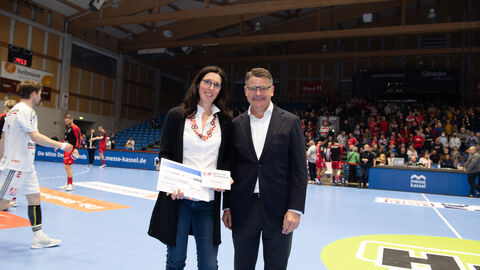 Ministerpräsident Boris Rhein steht auf dem Spielfeld der MT Melsungen. Die Ehrenamtliche Carolin Goujard hat von ihm eine Auszeichnung überreicht bekommen. Diese hält sie in der Hand.