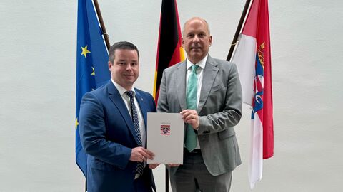 Andreas Hofmeister (links) mit Innenminister Poseck (rechts). Die beiden stehen nebeneinander und halten gemeinsam ein Dokument in den Händen. Im Hintergrund sind die Flaggen des Landes Hessen, Deutschlands und der EU zu sehen.